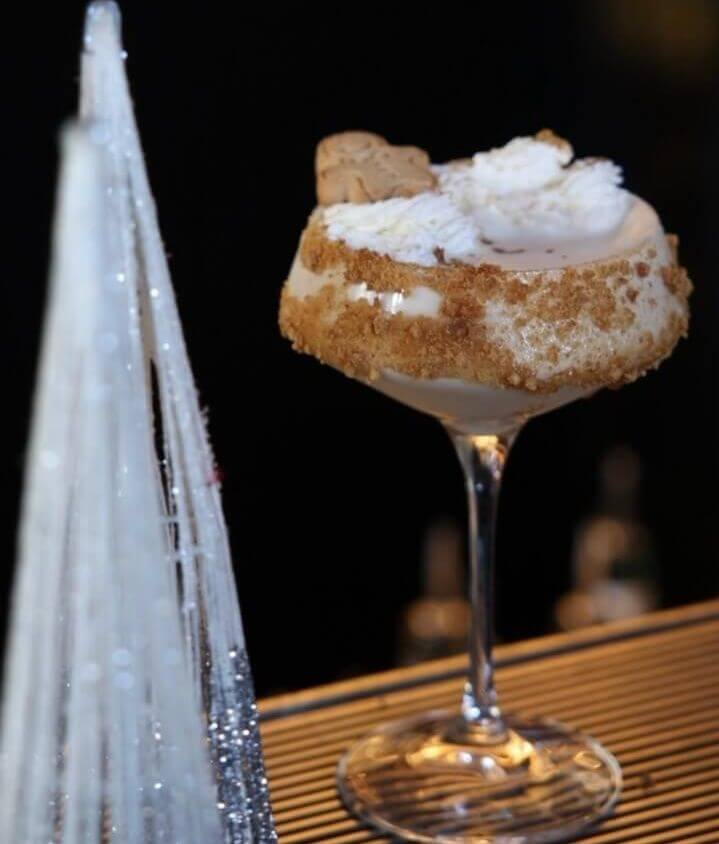 A martini glass filled with a beige coloured cocktail, topped with a gingerbread man cookie and cream